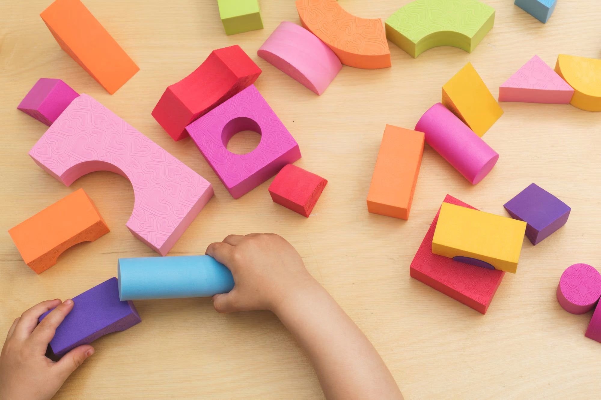Colorful foam blocks on a wooden surface with a child's hand interacting with them.